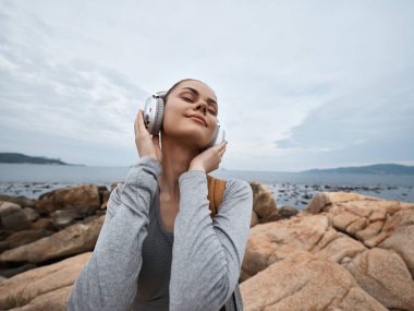 Günlük kıyafetli genç bir kadın, kayalık bir plajda kulaklıkla müzik dinliyor. Güzel sahil manzarasından zevk alırken neşe ve sükunet ifade ediyor.