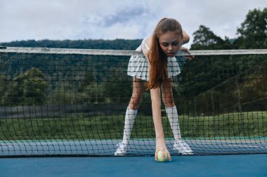 Sahada tenis topunu geri alan etekli genç bayan tenisçi, yeşil doğal zemin, somut kararlılık ve atletizm.