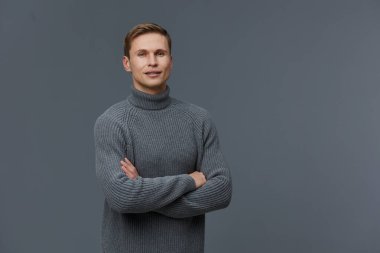 Gri örgü süveter giyen, kollarını çapraz bağlamış beyaz bir adam tarafsız gri bir arka planda kameraya güvenle bakıyor. Görüntü sakin ve profesyonel bir havayı yansıtıyor.