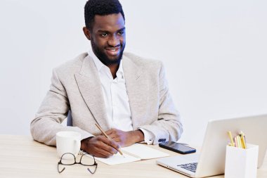 Modern ofis ortamında dizüstü bilgisayarı, defteri, yazma araçları, stüdyo ayarları, parlak ve davetkar bir atmosferde çalışarak resmi giyinmiş neşeli genç bir zenci.