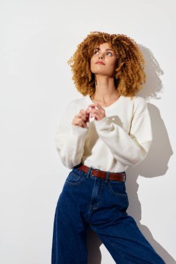 Beyaz kazak ve kot pantolon giymiş kıvırcık saçlı şık bir kadın temiz beyaz bir zemine karşı poz veriyor modern moda estetiğini ve günümüzün yaşam tarzını temsil eden sıradan bir havayı sergiliyor.