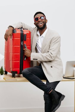 Çağdaş bir çalışma alanında kırmızı bavula yaslanmış koyu tenli ve son moda güneş gözlüklü mutlu bir adam. Açık renk ceket ve beyaz gömlek giyiyordu. Seyahat konusunda kendine güven ve heyecan duyuyordu.