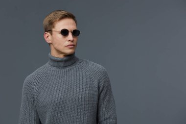 Gri balıkçı yaka kazak ve yuvarlak güneş gözlüğü takmış sarı saçlı, şık bir gencin portresi. Tarz ve güveni vurguluyor, sıradan moda.