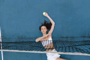 Enerjik bayan atlet şık beyaz spor kıyafetleriyle tenis topuna servis yapıyor, canlı mavi bir zemin üzerine kurulmuş, spor, fitness ve sağlıklı yaşam tarzı temaları için mükemmel.