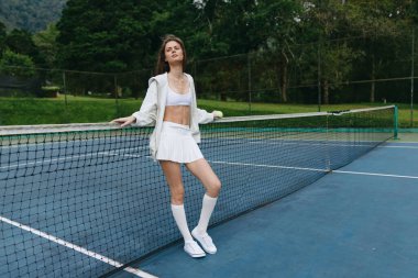 Kendine güvenen, şık beyaz spor elbiseli genç bir kadın tenis kortunda duruyor, sağlıklı bir yaşam tarzını, sporu ve kendini adamışlığı temsil ediyor.