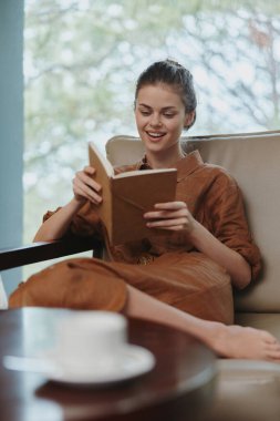 Kahverengi elbiseli genç bir kadın evde kitap okurken gülümsüyor. Kapalı mekandaki rahat ortam yumuşak doğal ışık, rahat bir atmosfer ve masada bir fincan çay içeriyor.