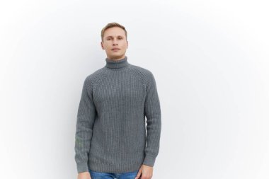Açık tenli, kısa sarı saçlı, mavi gözlü, kalın gri balıkçı yaka kazak giyen, beyaz arka planlı, minimalist bir stüdyoda güvenle poz veren bir adam. Moda için mükemmel, günlük kıyafet