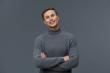 Samimi gri balıkçı yaka kazak giyen mutlu genç adam kollarını kavuşturup kameraya içten bir gülümsemeyle poz veriyor ve tarafsız gri bir arkaplanın önünde duruyor. Kavram: kendinden emin, gündelik tarz