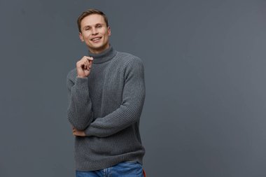 Gri balıkçı yaka kazak ve mavi kot pantolon giymiş gülümseyen genç beyaz adam gri arka planda tek başına duruyor. Mutlu, pozitif duygu, günlük tarz dijital göçebe kavramının yanında el ele tutuşun.