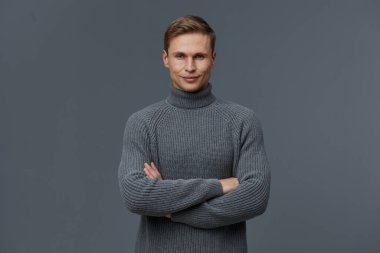 Gri boğazlı kazak giyen gülümseyen adam kolları çapraz duran ve koyu gri arka planda kameraya bakan. Dijital göçebe görünümlü kendinden emin beyaz bir adamın portresi.