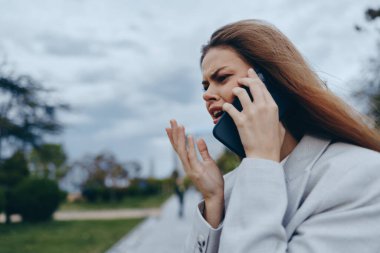 Profesyonel bir iş kadını bir telefon görüşmesi yapıyor. Bulutlu gökyüzünün altında doğal ve açık bir ortamda hayal kırıklığı ve kararlılık duygularını gösteriyor..