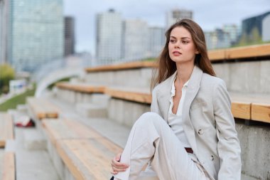 Hafif takım elbiseli zarif bir kadın modern şehir ortamında ahşap banklarda oturuyor, düşünceli bir ifade sergiliyor, moda tarzını psikolojik konsept ve sonbahar havasıyla harmanlıyor..