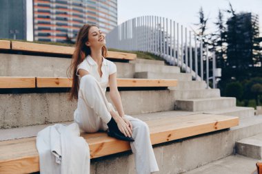 Beyaz elbiseli genç bir kadın ahşap basamaklarda oturuyor neşeli bir gülümseme, şehirli arka planı, sıradan moda ve bahar havası, rahat ve mutlu duyguları yakalıyor..