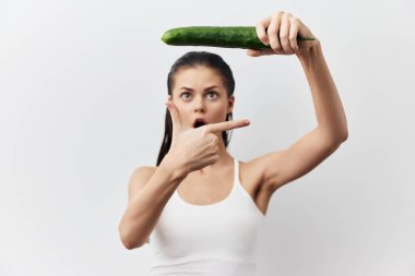 Şok olmuş bir kadın, parmağını salatalığı başının üstünde tutarken, şaşkınlık ifadesi sergiliyor, hafif nötr bir arka plana yaslanıyor, mizahı ve yaratıcılığı yakalıyor.