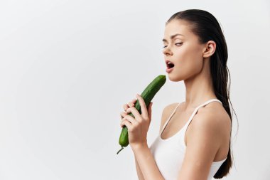 Islak saçlı ve kendine güveni olan genç bir kadın dudaklarının yanında bir salatalık tutuyor. Tarafsız beyaz arka plan yemek ve şehvet kavramını vurguluyor.