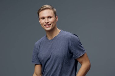 Kısa açık kahverengi saçlı, sıradan mavi tişörtlü neşeli genç bir adam. Sade gri arka planda dikilirken sıcak bir şekilde gülümsüyor. Stüdyo portresi pozitif ruh hali ve arkadaşlığı yansıtıyor.