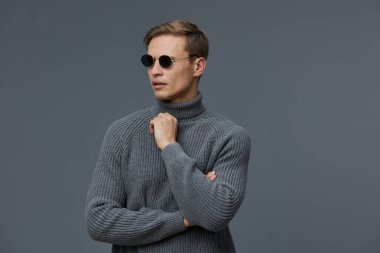 Güneş gözlüklü ve gri balıkçı yaka kazaklı düşünceli bir adam gri stüdyo arka planına karşı kollarını kavuşturmuş dijital göçebe yaşam tarzını ve uzaktan çalışma esnekliğini sembolize eden yanlara bakıyor.