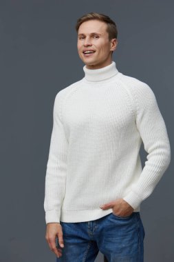 Kısa kahverengi saçlı neşeli genç bir adam, rahat beyaz bir balıkçı yaka ve sıradan bir kot pantolon giymiş, kendine güvenerek duruyor ve gülümsüyor, tarafsız gri bir arka plana karşı rahat bir poz veriyor. Kavram