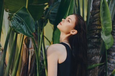 Palmiye ağaçlarının önünde duran uzun siyah saçlı kadın güneşli bir günde gökyüzüne bakıyor.