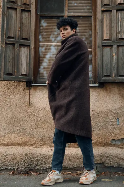Kahverengi paltolu ve spor ayakkabılı şık genç adam eski binanın önünde elleri ceplerinde duruyor..