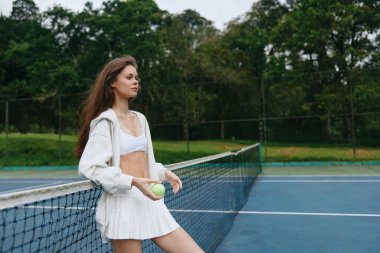 Beyaz etek ve tenis eteği giyen genç atletik model, güvenli bir şekilde bir tenis ağına yaslanıyor, doğa ile çevrili bu canlı görüntü fitness, spor ve canlılığı vurguluyor.