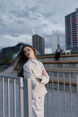 Açık renk ceketli kadın şehir binaları ve bulutlu gökyüzü arka planı ile güvenli bir şekilde dışarıda duruyor. Sıradan moda tarzını, modern şehir yaşam tarzını ve kasvetli atmosferini somutlaştırıyor.