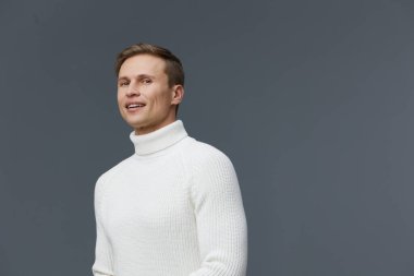 Kısa kahverengi saçlı, rahat, beyaz balıkçı yaka kazaklı, güler yüzlü, güler yüzlü, basit, gri bir arka planda duran neşeli bir genç. Kavram: Modern gündelik biçim.