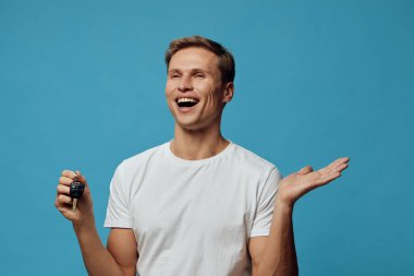 Günlük kıyafetleri ve kısa kahverengi saçlarıyla gülümseyen genç adam mutluluk ve heyecanı ifade ediyor, bir araba anahtarı tutuyor, hayat tarzı ve otomotiv temaları için canlı mavi bir arka planda. Kavram: sevincin