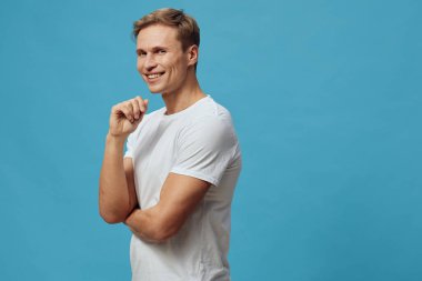 Gülümseyen, beyaz tişörtlü, kolları çapraz duran genç beyaz bir adam mavi arka planda izole bir şekilde poz veriyor. Konsept: Uzak çalışma ile dijital göçebe yaşam tarzı