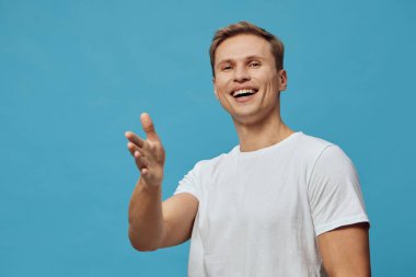 Beyaz tişörtlü, gülümseyen bir adam açık mavi, pürüzsüz arka planda doğal neşeli bir ifadeyle elini öne uzatıyor. Sosyal etkileşim ve dijital etkileşimi temsil eden günlük tarz ve dostça bir jest