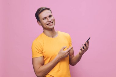 Kısa saçlı, sarı tişörtlü neşeli bir kadın akıllı telefonunu kullanmaktan zevk alıyor. Cihazla uğraşırken geniş bir şekilde gülümsüyor, düz pembe bir arka plan ile canlı ve pozitif bir ortam yaratıyor.