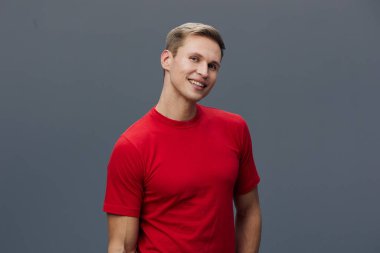 Sarı saçlı, parlak tebessümlü, kırmızı günlük tişörtlü, sade gri arka planda kendine güvenen genç bir adam. Stüdyo çekimi neşeli ifadeler yakalar, rahatlatıcı atmosfer