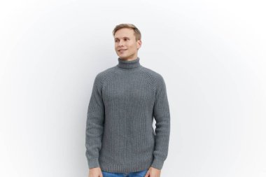 Gri balıkçı yaka kazaklı, gülümseyen adam beyaz arka planda tek başına dikiliyor ve yana bakıyor. Gündelik sonbahar kış kıyafetleri, rahat duruş, ruh sağlığı ve sakince ifade edilen aletler teması