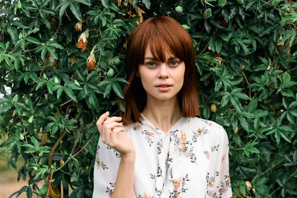 Canlı kızıl saçlı kadın yemyeşil çalıların önünde yapraklarla dikiliyor.