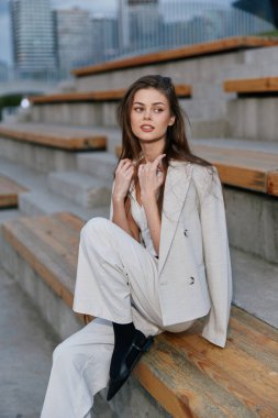 Açık renk ceketli ve pantolonlu zarif bir kadın şehir ortamında ahşap merdivenlerde oturuyor. Düşünceli bir ifade, moda tarzı, sonbahar sezonu, moda kadını.