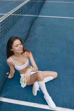 Beyaz spor giyimli genç bir bayan tenis topuyla tenis kortunda otururken gülümsüyor, canlı mavi bir arka plana karşı neşeli ve enerjik bir spor tarzı sergiliyor.