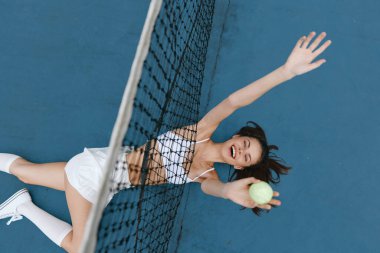 Canlı, genç bir kadın, spor giysileri içinde, mavi kortta tenis oynamaktan zevk alıyor, topa uzanırken heyecanını dile getiriyor, atletizm ve neşenin özünü yakalıyor.