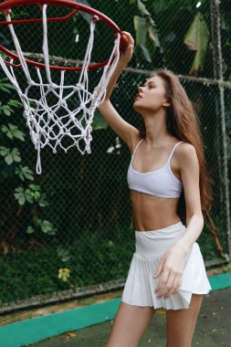 Beyaz ekin bluz ve etek giyen sportif bir kadın model. Basket potasına uzanırken, sağlıklı ve formda olmak için ideal, canlı bir ortamda kararlılığını gösteriyor.