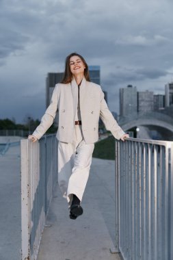Bej takım elbiseli kendine güvenen genç bir kadın şehir ortamında modern binalara ve bulutlu gökyüzüne neşeyle gülümsüyor. Moda dünyası kavramı.