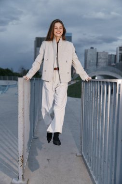 Açık renkli iş kıyafeti giymiş kendine güvenen bir kadın, gülümseyen ve açık havada şehrin siluetine karşı poz veren, modern profesyonelliği ve güçlendirmeyi somutlaştıran.