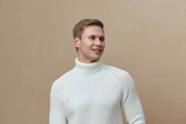 Beyaz örgülü balıkçı yaka kazak giyen, gülümseyen, beyaz tenli genç bir adam olumlu duygularla yan tarafa bakıyor. Temiz bej arka plan gelişigüzel kış modası ve zihinsel sağlık araçları kavramını vurguluyor