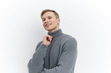 Sade gri balıkçı yaka kazak giyen mutlu bir adam, gülümseyen ve düşünceli bir şekilde başka yöne bakan, düz beyaz bir arka plan karşısında duran. Görüntü rahatlığı, sıcaklığı ve rahatlığı vurguluyor.