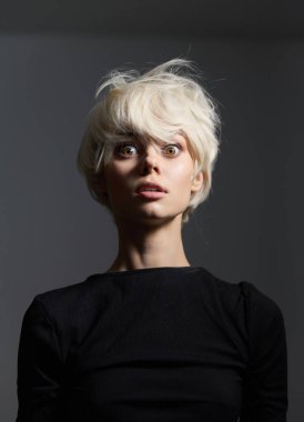 Kısa sarı saçları ve geniş gözleri olan, sade siyah bir gömlek giyen genç bir kadın, yumuşak gri bir arka plana karşı yoğun bir sürpriz anı yakalar..