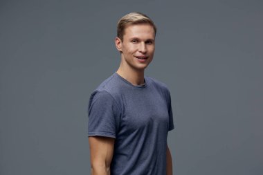 Kısa sarı saçlı, sıradan mavi tişörtlü, kendine güvenen mutlu genç bir adam, tarafsız gri bir stüdyo arkaplanına poz verdi. Kavram: pozitif tutum ve rahat biçim