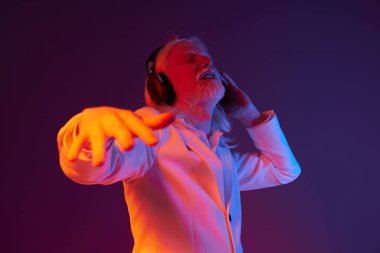 Beyaz ceketli yaşlı bir adam, kulaklık takarken müziğe dalmış, canlı mor ve turuncu arka plan dinamik havayı güçlendiriyor, neşe ve ritmi dışavurumcu vücut dili ile gösteriyor.