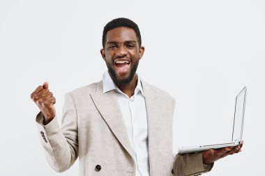 Koyu tenli, bej ceketli ve beyaz gömlekli mutlu bir adam elinde bir dizüstü bilgisayar tutarak heyecanını dile getiriyor. Düz beyaz bir arkaplan karşısında duruyor. Kavram: başarı ve coşku.