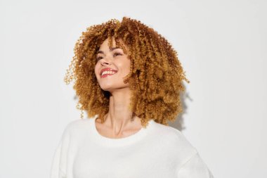 Şehvetli kıvırcık saçlı mutlu bir kadın, sıcak bir gülümseme sergiliyor, basit beyaz bir arka plana karşı duruyor, doğal güzelliğini arttırıyor ve bu parlak moda portrelerinde kendine güven ve neşe yayıyor.