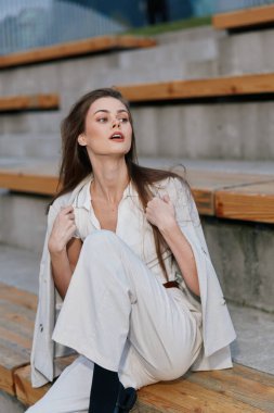 Zarif beyaz elbiseli, kendine güvenen genç beyaz kadın ve ceket tarafsız gri şehir ortamında ahşap merdivenlerde oturuyor, sonbahar moda tarzını sakin duygularla yakalıyor..