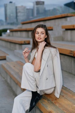 Açık bej takım elbiseli genç bir kadın ahşap şehir merdivenlerinde oturuyor. Düşünceli bir ifadeyle, moda tarzını ve bahar şehri ortamında günlük iş kıyafetlerini sergiliyor.