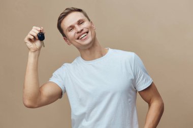 Kısa kahverengi saçlı neşeli bir adam, açık mavi tişört giyiyor, temiz anahtarlar sergiliyor, mutlu bir ifade sergiliyor, bej renkli stüdyo geçmişine karşı poz veriyor, araba sahipliğini kutluyor, pozitif.